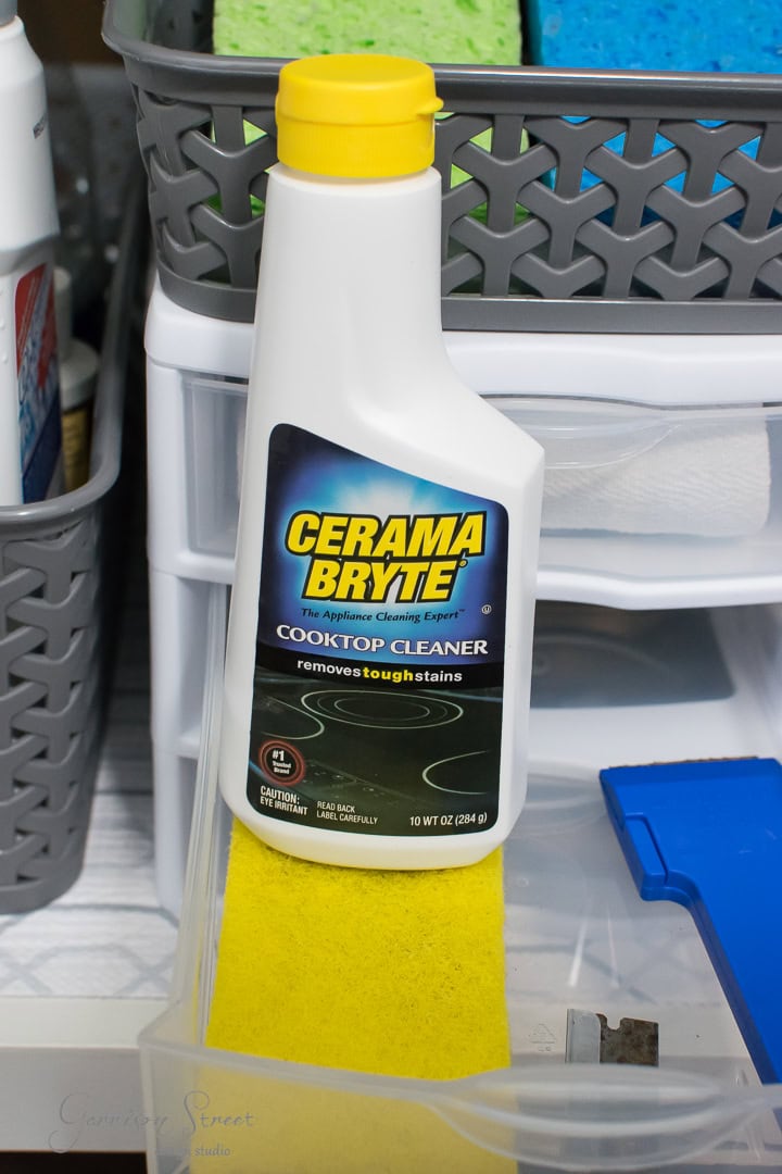 A bottle of Cerama Bryte Cooktop Cleaner rests on a yellow sponge, surrounded by cleaning supplies and plastic storage bins for perfect organization. This cleaner is essential for removing tough stains from your kitchen sink or cooktops.
