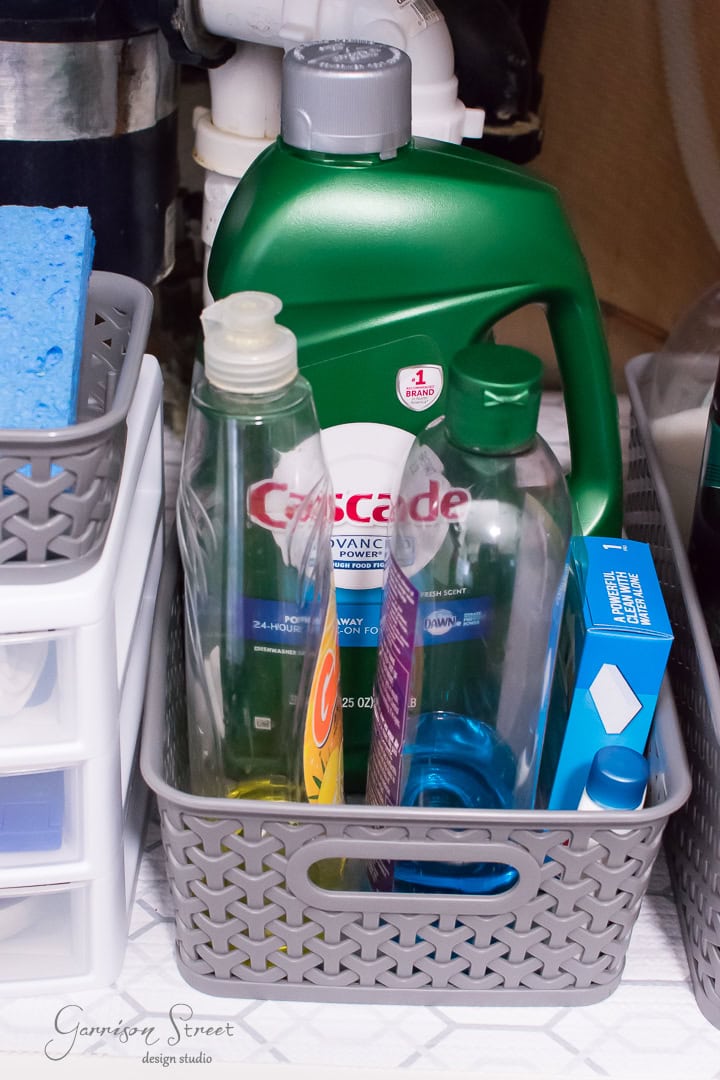 A storage basket under the kitchen sink holds cleaning supplies: a large green bottle of Cascade detergent, a yellow dish soap bottle, and a blue box. Nearby on the shelf is a blue sponge. A drawer unit is partially visible to the left, adding an element of organization to the space.