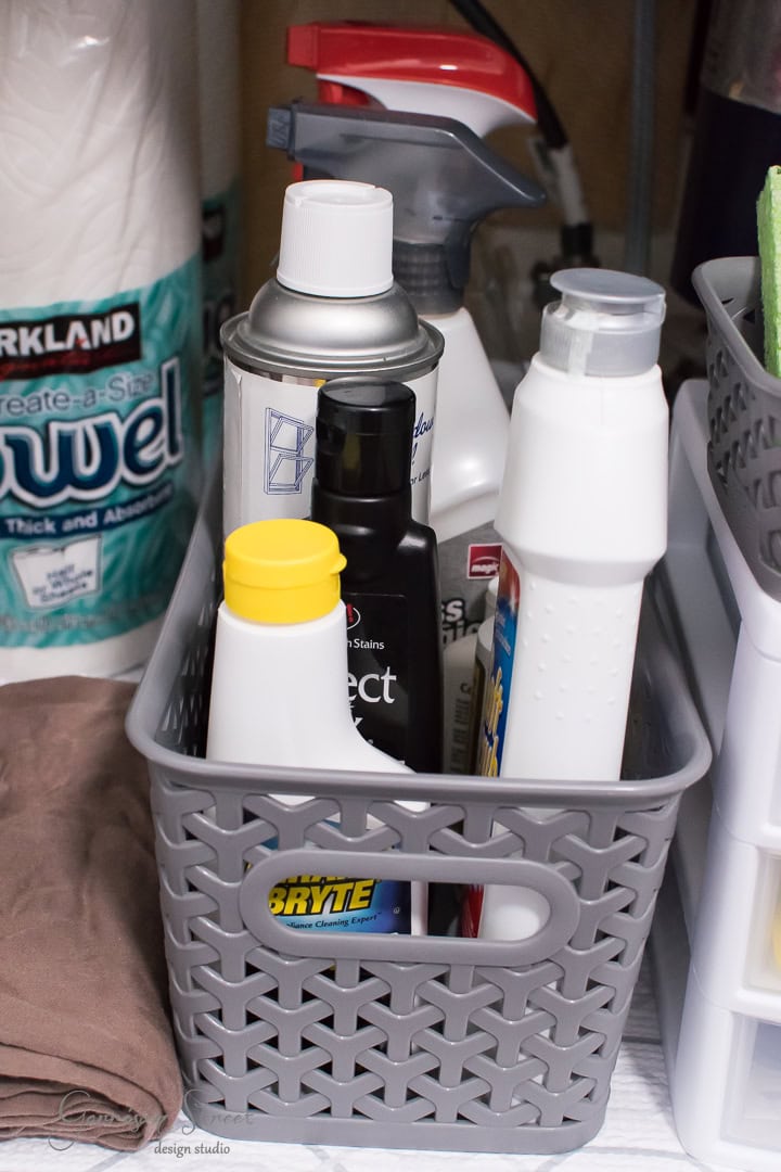 A gray basket filled with various cleaning supplies, including spray bottles and cans, sits on a shelf. Nearby, paper towels and folded cloths are organized neatly in a utility area by the kitchen sink to enhance organization.