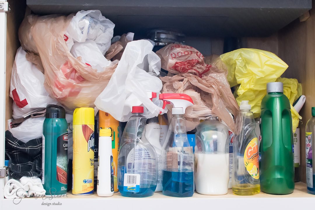 A cluttered cabinet under the kitchen sink houses various cleaning supplies, from plastic spray bottles to aerosol cans and plastic bags. The random stacking of items calls out for organization, creating a disorganized appearance.