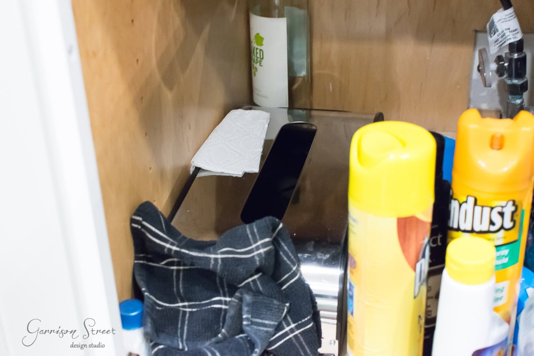 A well-organized kitchen cabinet holds various items: a bottle on a shelf, a paper towel neatly placed, a small black appliance on the lower shelf, alongside a yellow can of spray, dusting spray for effortless cleaning, and a dark cloth—everything within easy reach by the kitchen sink.