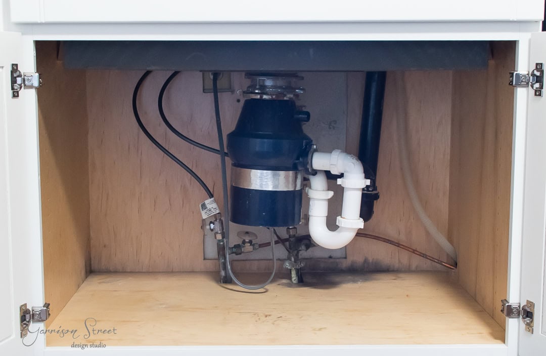 The image shows the underside of a well-organized kitchen sink cabinet, revealing plumbing pipes and a garbage disposal unit connected to a white P-trap pipe. The wooden walls of the cabinet neatly contain some visible electrical wires.