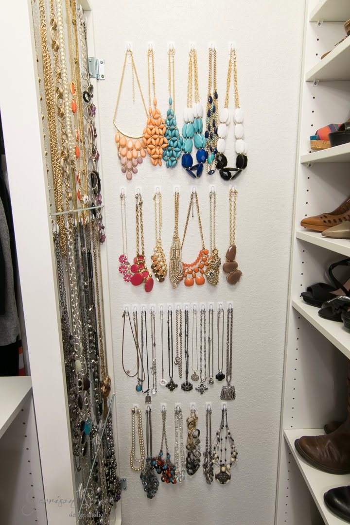A neatly organized jewelry closet featuring multiple necklaces hanging in rows by color and style, with a variety of beads, chains, and pendants. An IKEA mirror adds elegance, while shelves on the side showcase neatly arranged shoes.