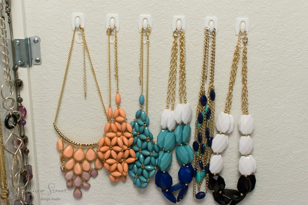 Assorted colorful necklaces with large beads hang on white hooks against a textured wall beside an elegant mirror. The necklaces, reminiscent of an IKEA display, feature gold chains and beads in shades of pink, orange, turquoise, blue, white, and black.