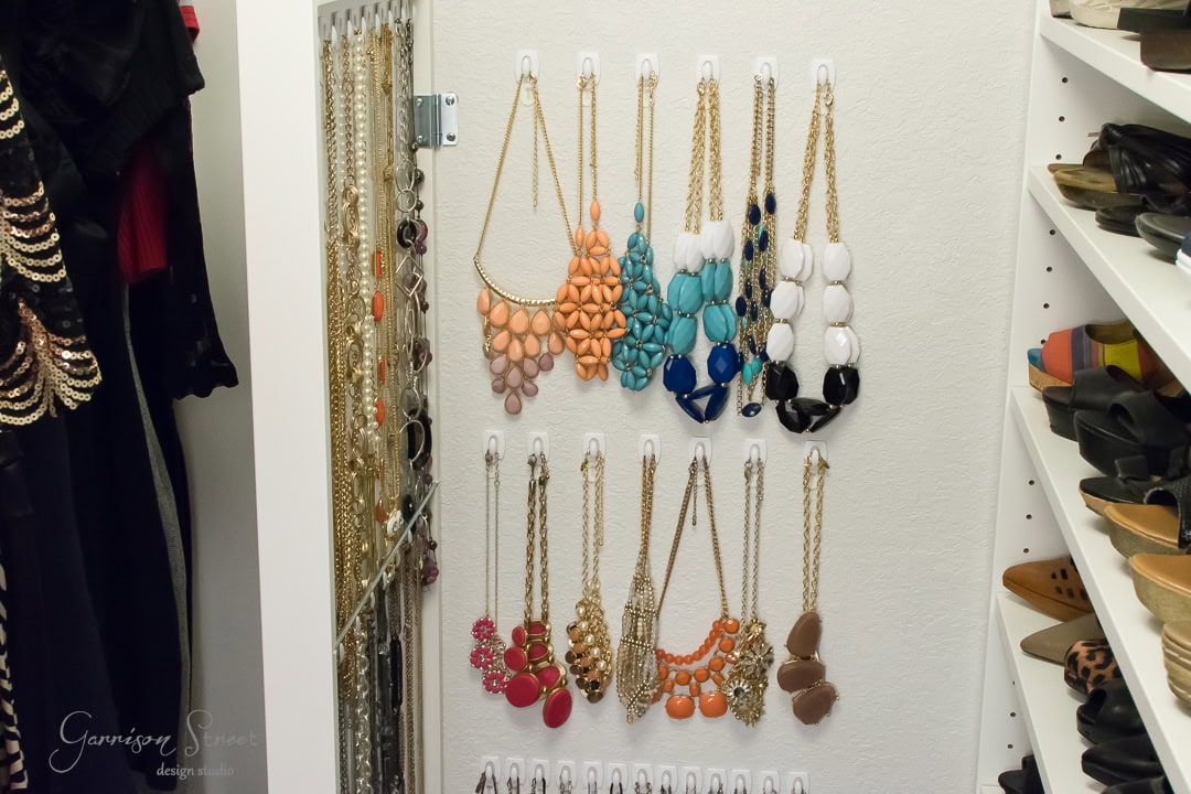 A closet corner featuring an IKEA jewelry organizer with multiple necklaces, including colorful beads and gold chains, neatly hung on hooks. A nearby mirror reflects shelves displaying assorted shoes and clothing.