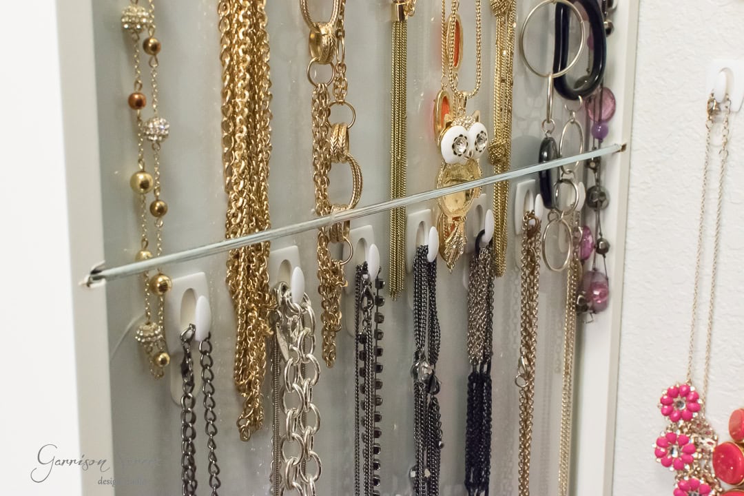 A close-up of a jewelry organizer from IKEA reveals an array of necklaces and bracelets elegantly hanging. The collection boasts gold, silver, and black chains, with decorative beads and charms. A few colorful earrings glisten beside a small mirror reflecting the dazzling display.
