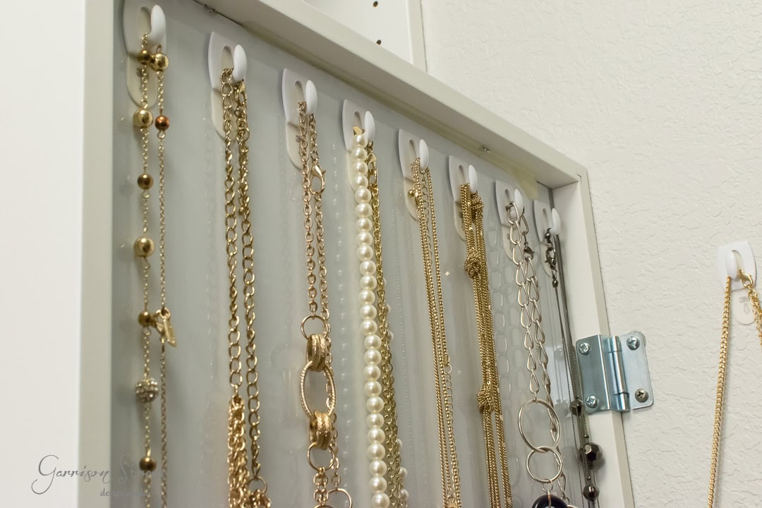 A sleek white cabinet, reminiscent of IKEA's minimalist style, hangs on the wall as a jewelry organizer. Its mirrored door opens to reveal neatly displayed necklaces, from gold chains to pearls, elegantly arranged on hooks inside.