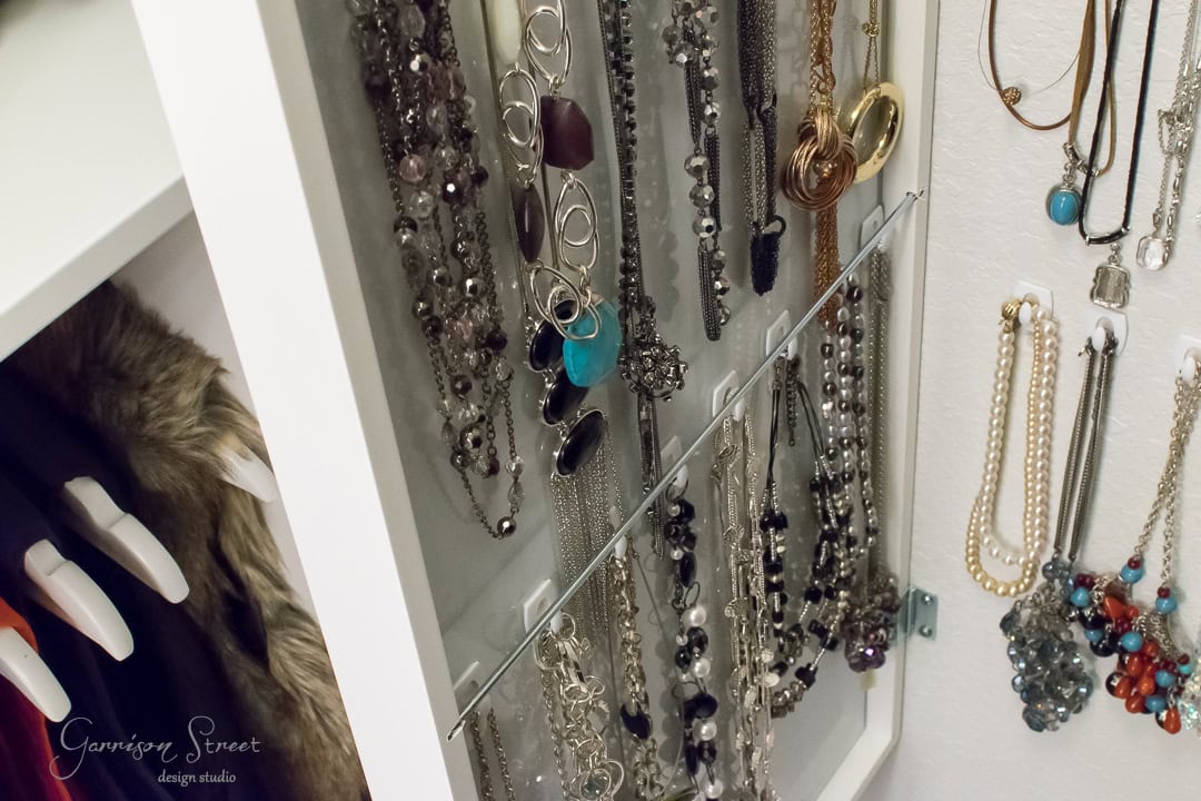 Inside a white cabinet, an IKEA mirror reflects a jewelry organizer displaying various necklaces—silver chains, beaded pieces, and pendants—all neatly hung. Some feature blue accents while fur-lined jackets hang to the side, adding charm to this elegant setup.