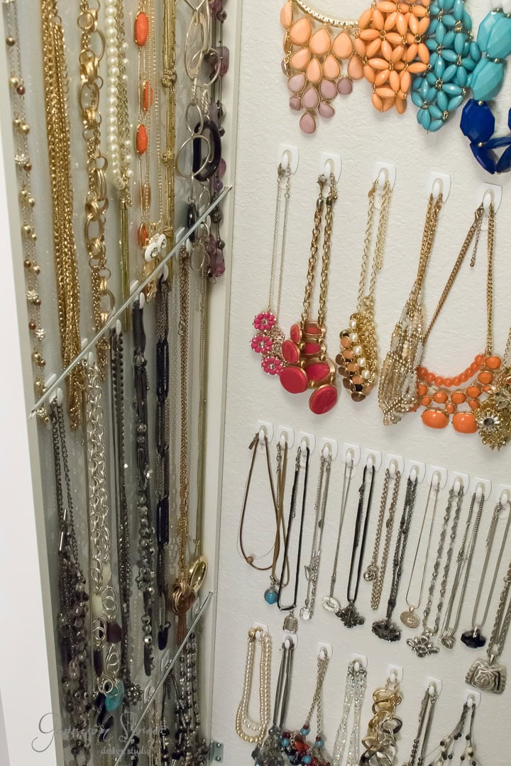 A neatly organized jewelry display features various necklaces and bracelets hanging on hooks. The collection, including gold, silver, and colorful beaded pieces, is elegantly arranged on an IKEA wall-mounted organizer with a nearby mirror to perfectly complement the presentation.