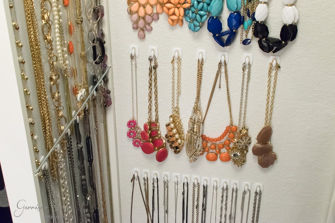 A collection of necklaces and earrings organized on an IKEA wall display. The necklaces feature various designs and colors, including gold, pink, orange, and brown. Earrings are neatly hung above, their vibrant styles reflecting beautifully in an adjacent mirror.
