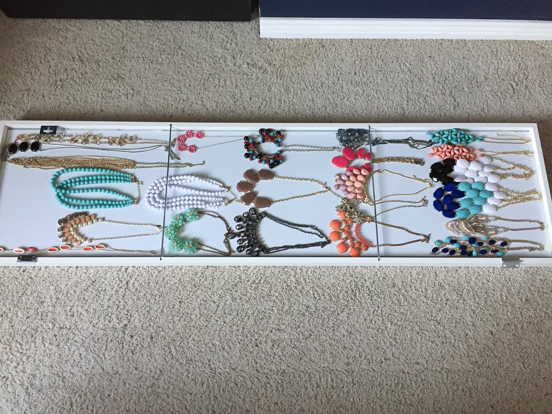 A variety of colorful necklaces are neatly arranged on a white tray placed on a beige carpet. The necklaces, paired stylishly with an IKEA mirror, feature beads and pendants in vibrant hues like teal, pink, orange, black, and white, organized meticulously in rows.