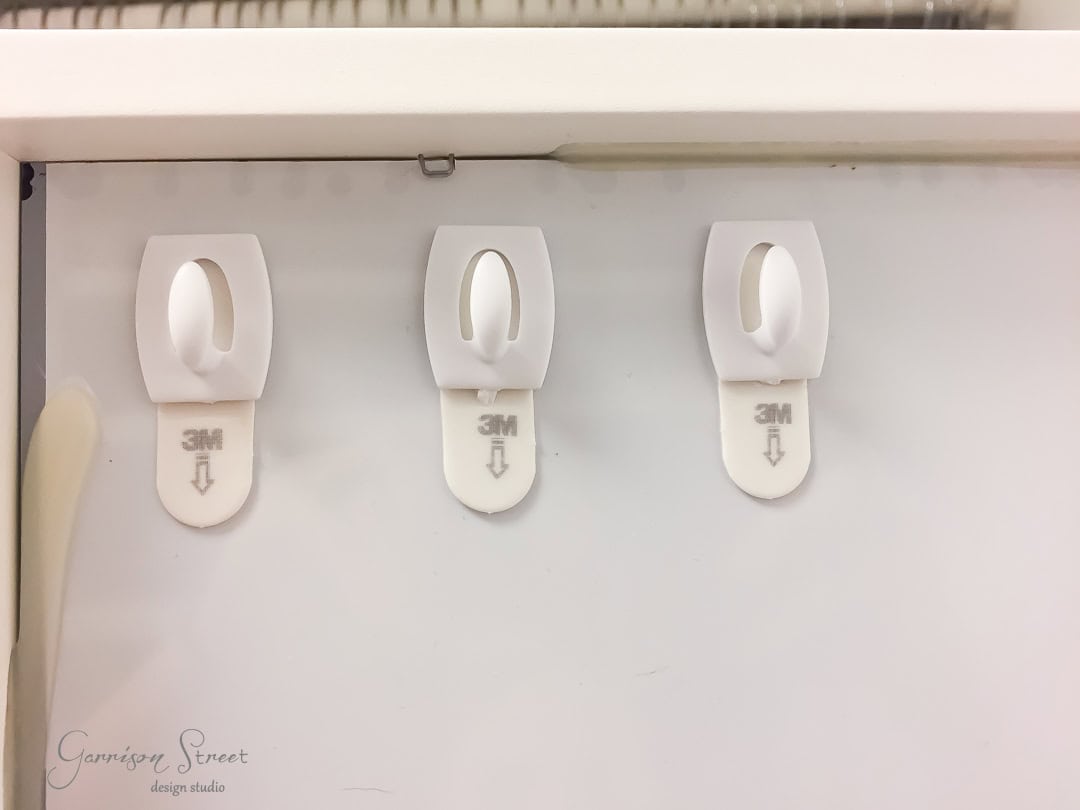Three white adhesive wall hooks, reminiscent of Ikea's minimalist design, are mounted on a vertical surface. Each hook displays the brand name and a downward arrow indicating proper usage. Positioned near a mirror, their sleek simplicity complements the plain, light-colored backdrop perfectly.