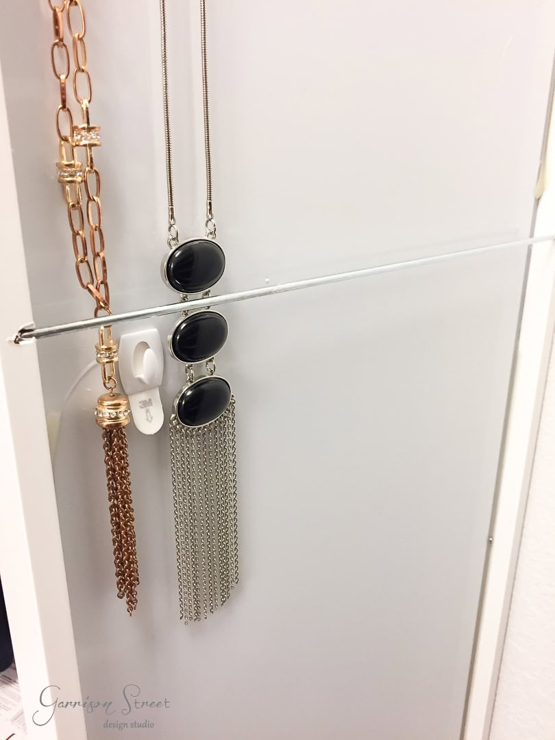 A sleek, white wall-mounted jewelry organizer from IKEA elegantly displays necklaces. A necklace with gold chains hangs on the left, while two with black pendants and silver fringes grace the right. A mirrored finish adds a touch of sophistication to this stylish display solution.