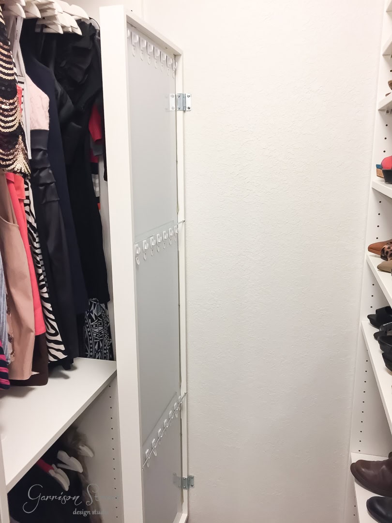 A narrow walk-in closet featuring neatly hung dresses and clothes on the left side is complemented by an IKEA shelf on the right, holding various shoes. A mirror enhances the well-organized space with a light-colored interior.