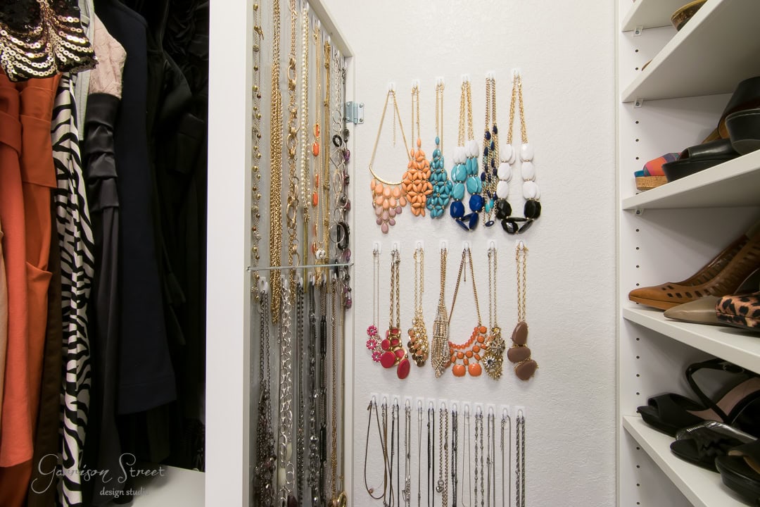 A well-organized closet with a section for necklaces hanging neatly on hooks. The collection includes various colorful beads and chains. An IKEA mirror reflects the space, while clothes hang to the left and shelves on the right store additional items.
