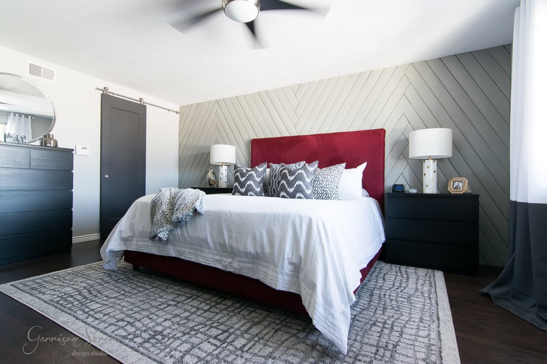 A modern bedroom features a large bed with a bold red headboard and white bedding, accented by patterned pillows. A geometric accent wall, two dark nightstands, and a dresser enhance the room's sleek design. A ceiling fan hangs above.