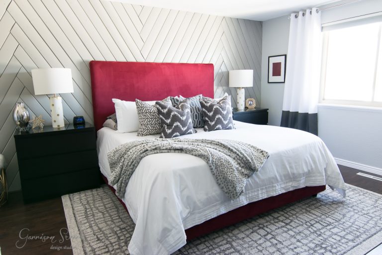 A modern bedroom with a large bed featuring a red headboard and white bedding. Two dark bedside tables with lamps are on either side. A geometric accent wall and a window with white curtains enhance the room's design.