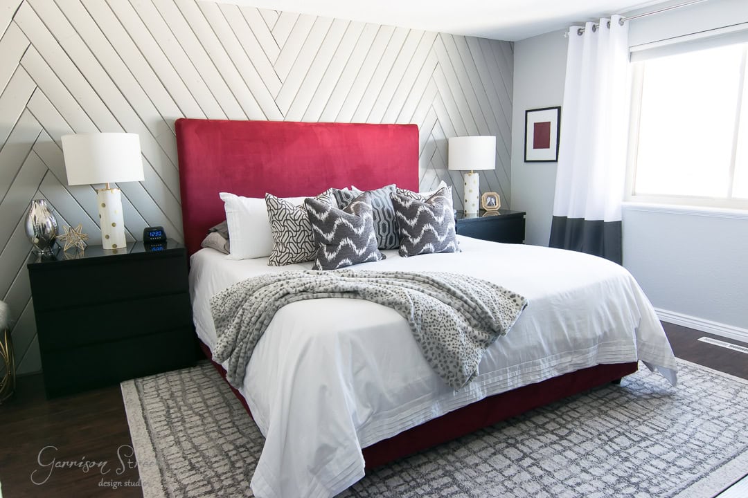 A modern bedroom with a large bed featuring a red headboard and white bedding. Two dark bedside tables with lamps are on either side. A geometric accent wall and a window with white curtains enhance the room's design.