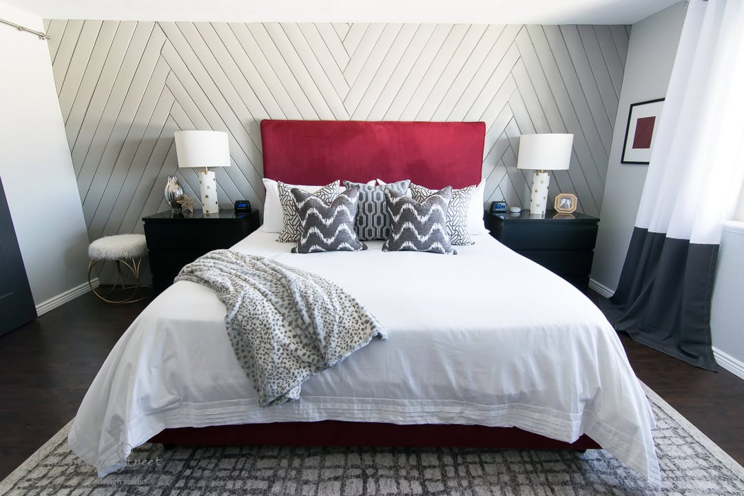 A modern bedroom with a red-upholstered headboard and white bedding featuring patterned pillows. A gray blanket is draped over the bed. There are black nightstands with lamps on each side, and a chevron-patterned accent wall behind. A rug covers the floor.