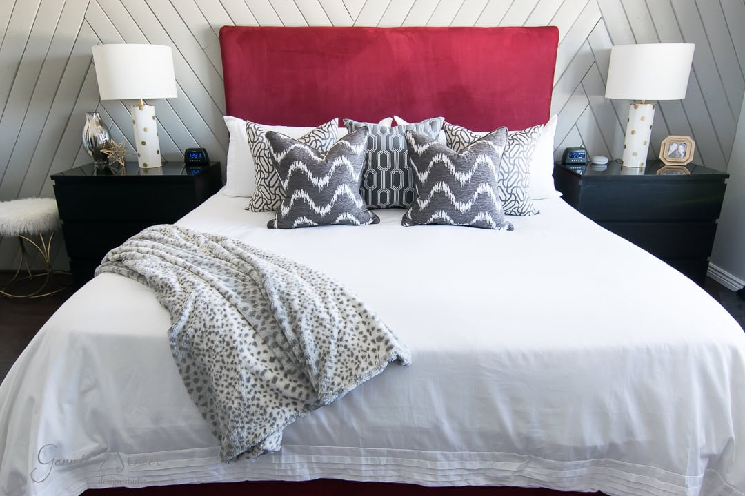 A bedroom with a red upholstered headboard and a neatly made bed. The bed has a white comforter, patterned throw pillows, and a spotted blanket. Two black nightstands with lamps and small decorative items sit on either side of the bed.