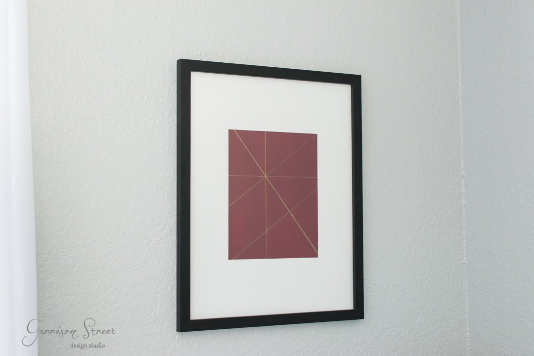 A framed abstract artwork hangs on a white wall. The piece features a maroon square with intersecting thin gold lines, creating geometric shapes. The frame is black with a white mat around the artwork.