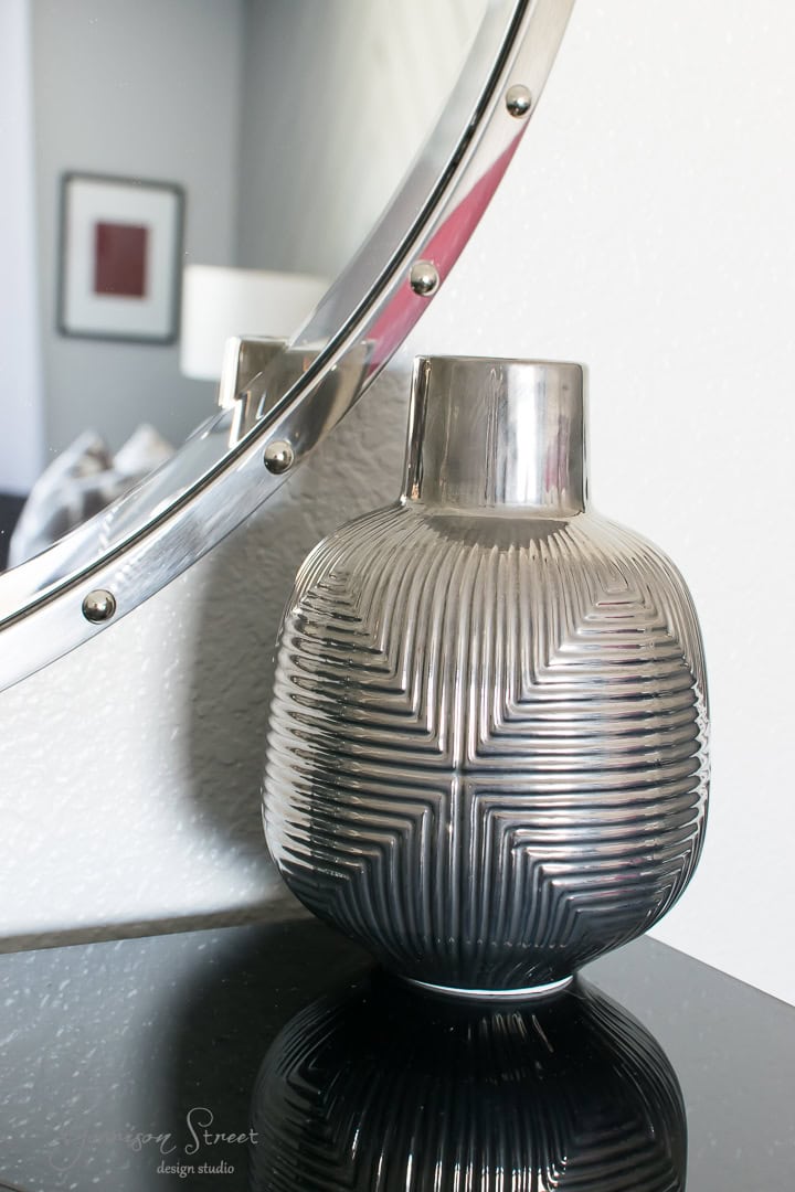 A metallic vase with a textured, geometric pattern sits on a black surface next to a round, silver-framed mirror. Soft light reflects off the smooth and ridged surfaces, highlighting the modern decor.