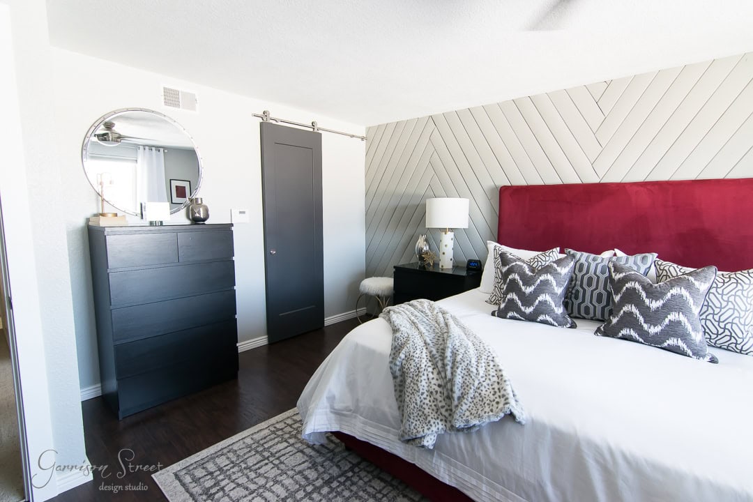 A modern bedroom with a red headboard, white bedding, and patterned pillows. A circular mirror hangs above a black dresser. A sliding barn door is on the side and the floor has a gray and white patterned rug. A lamp sits on a nightstand by the bed.