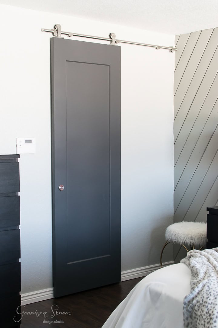 A sleek, dark gray sliding barn door on a silver track in a modern room with a textured wall and minimal decor. A fur-topped stool and a portion of a bed are visible, creating a cozy and stylish ambiance.