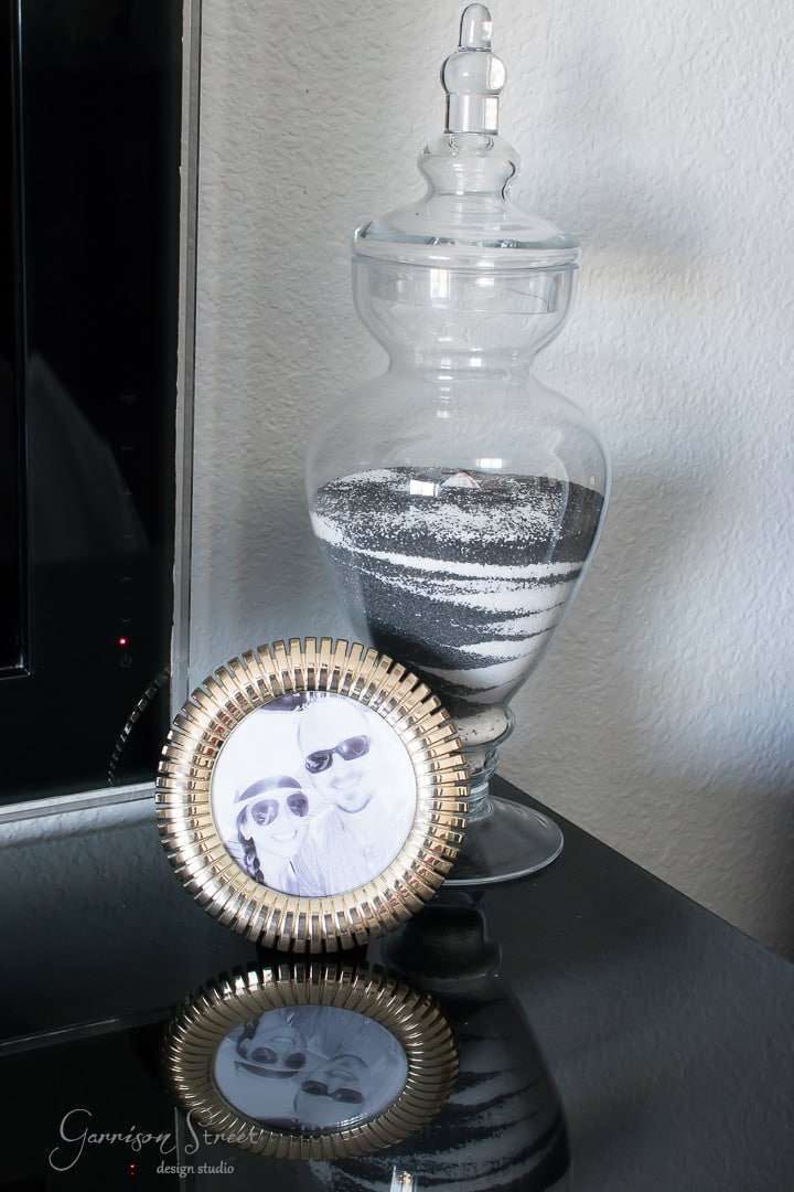 A decorative glass jar with layered black and white sand sits on a glossy surface. Beside it, there's a framed black and white photo of a couple wearing sunglasses, displayed in a gold-rimmed circular frame.