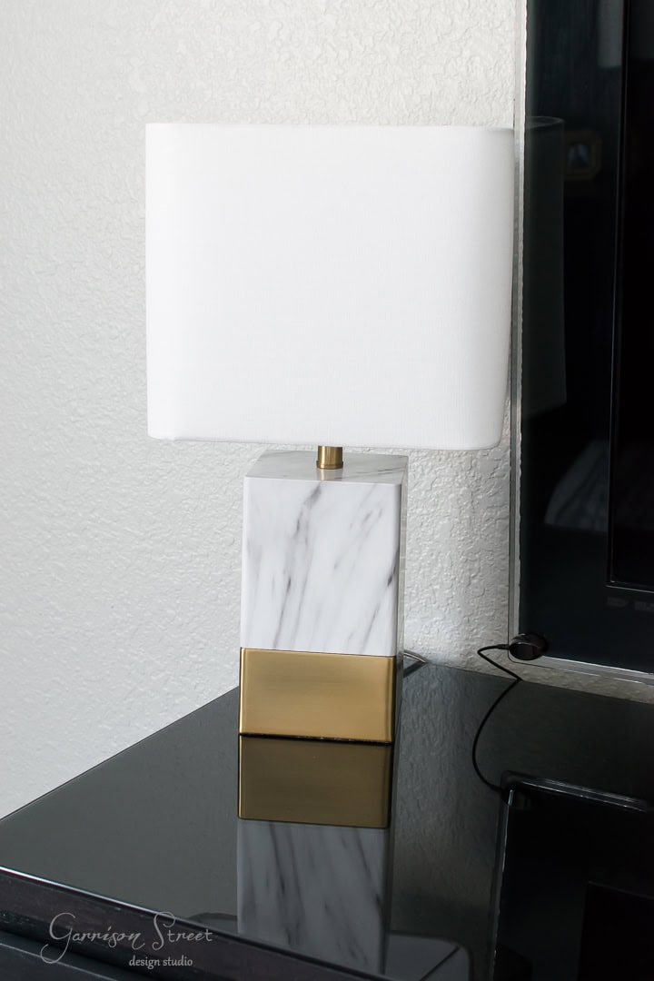 A modern table lamp with a square white shade and a marble and gold base sits on a glossy black surface next to a wall, with a plugged-in cord visible.