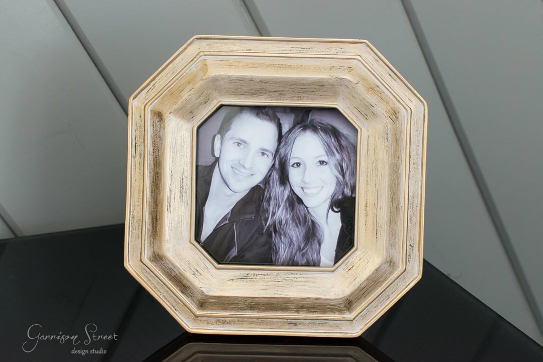 A black and white photograph of a smiling couple in a gold octagonal frame, placed on a black surface with a wooden paneled background.