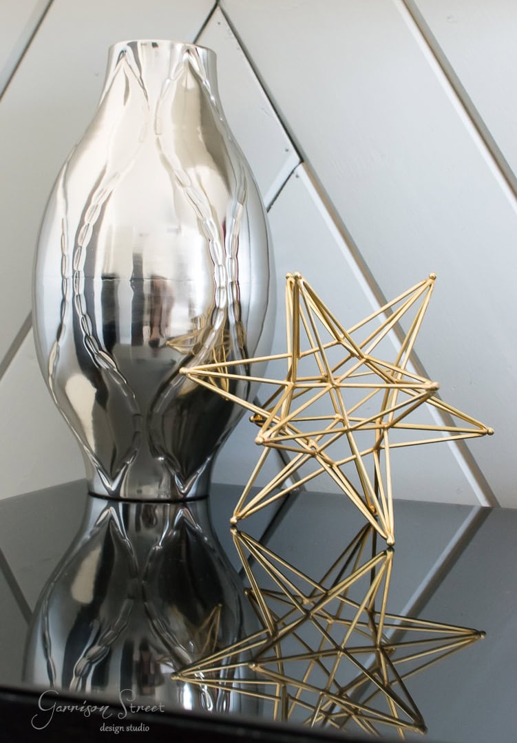 A reflective silver vase stands on a glossy black surface beside a geometric gold star sculpture. The objects and their reflections create a modern, elegant composition against a chevron-patterned wall in the background.