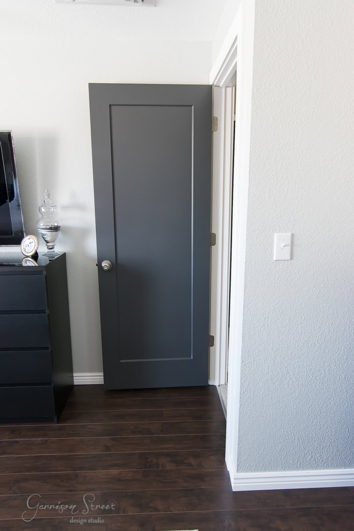 A dark gray door is partially open in a modern room with light gray walls and dark hardwood floors. A black dresser with a decorative item and a clock is nearby.