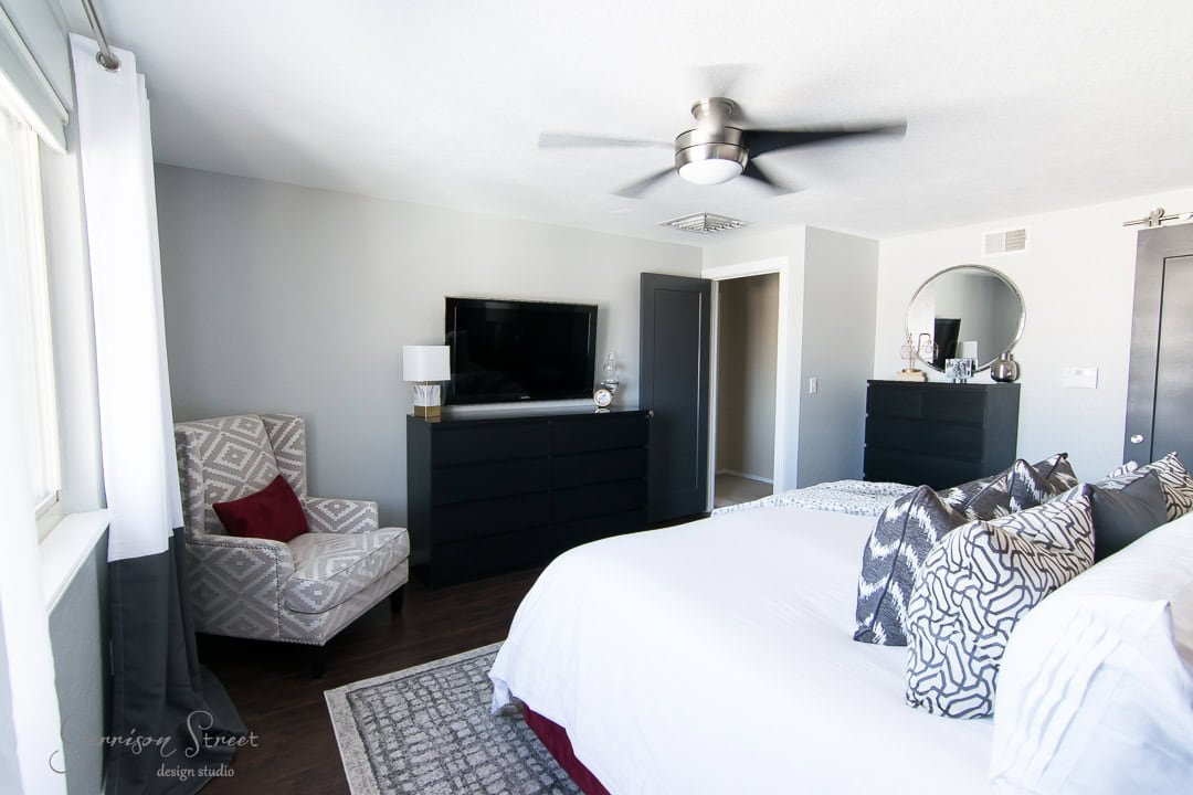 A modern bedroom with a large bed, patterned cushions, and a gray upholstered chair. A flat-screen TV sits on a black dresser, and there's a round mirror on another dresser. The room has a ceiling fan and a large window with curtains.