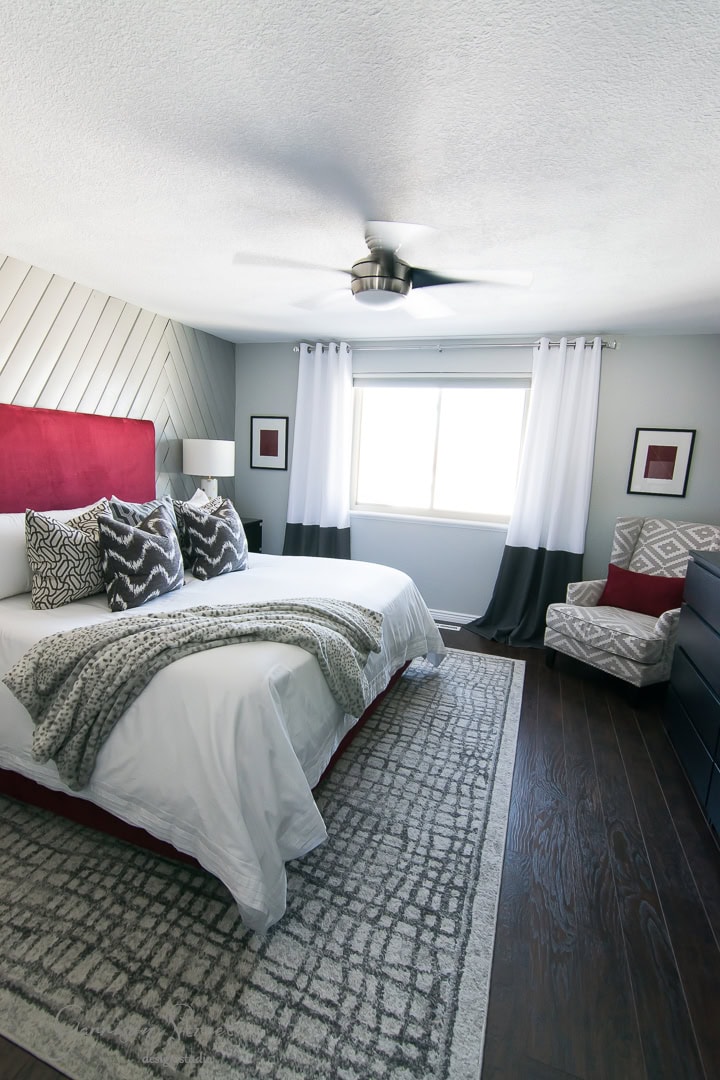 A modern bedroom showcases a large bed with red and gray accents, white and gray bedding, and geometric-patterned pillows. A cozy armchair near the window complements black and white curtains, while DIY art pieces add a personal touch to the decor.