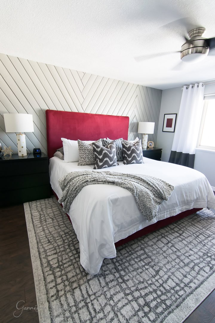 A modern bedroom with a red headboard and a chevron-patterned accent wall. The bed is adorned with white and gray bedding and decorative pillows. Black nightstands with lamps flank the bed. A ceiling fan and a window with white curtains are visible.