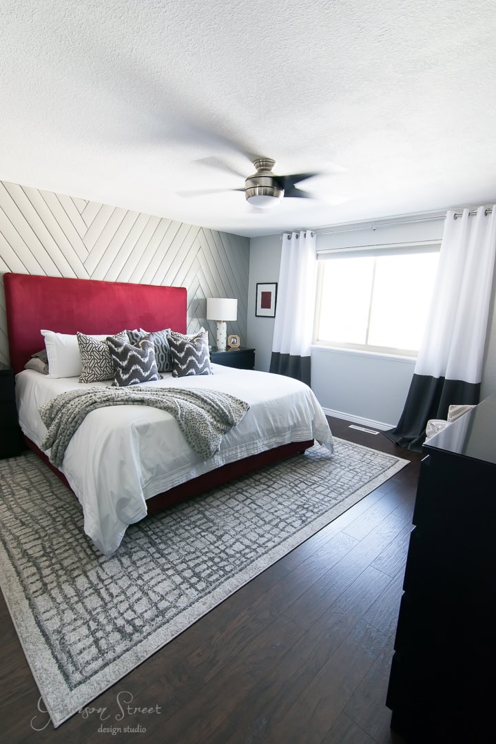 A modern bedroom with a red upholstered headboard, white bedding, and gray accent pillows. The walls feature a chevron pattern, and the room has a large window with white and gray curtains. There's a ceiling fan and a gray patterned rug on wooden flooring.