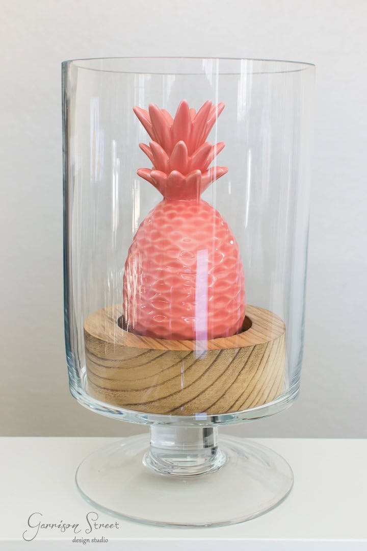 A pink ceramic pineapple sits inside a clear glass vase with a wooden base. The vase is placed on a white surface, and the background is a plain wall.