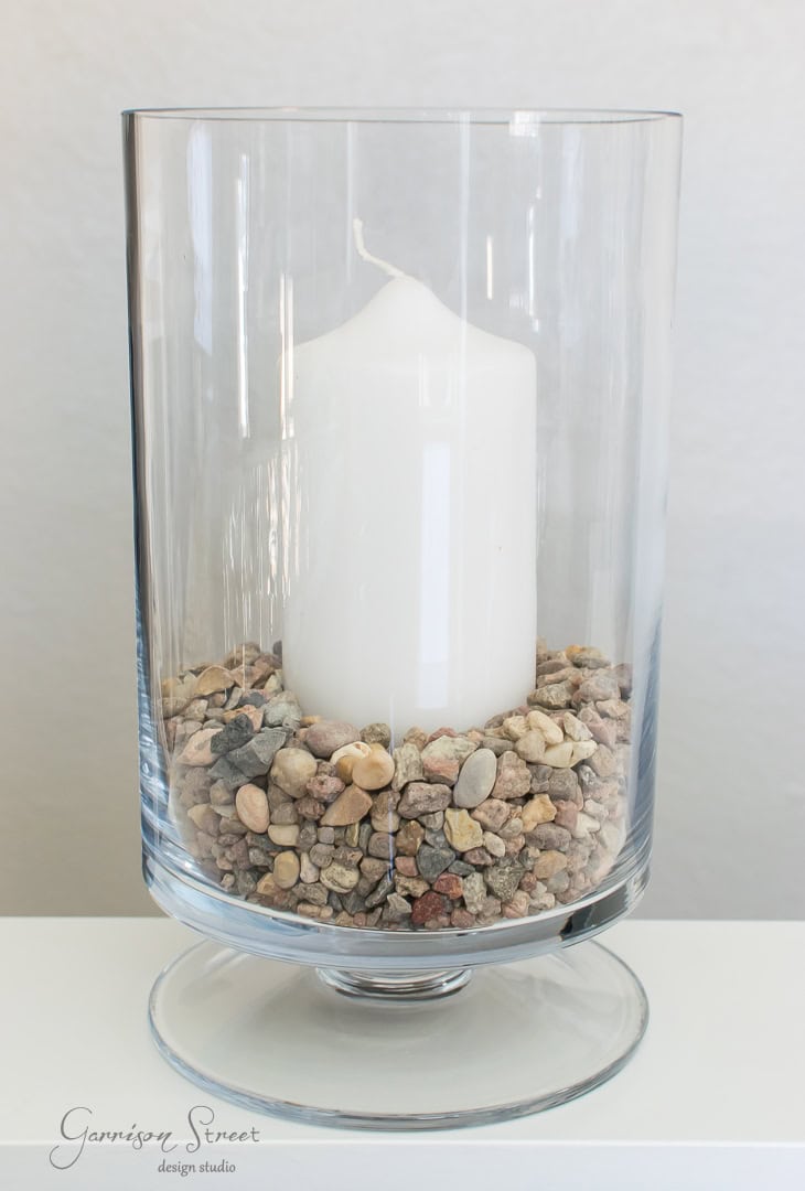A large white candle is placed inside a clear glass hurricane vase, surrounded by small multicolored pebbles. The vase sits on a light-colored surface against a plain background, creating a simple and elegant decorative piece.