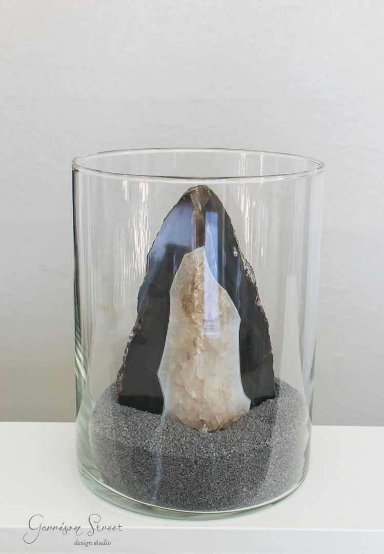 A decorative piece featuring a large crystal geode displayed vertically in a clear glass container filled with small gray pebbles at the base. The background is a plain white wall.
