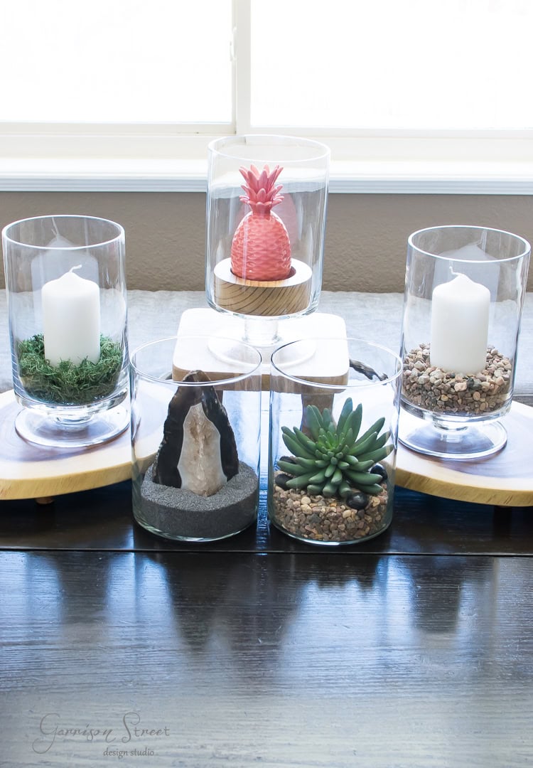 A collection of six glass containers on wooden stands, featuring candles, stones, moss, a small succulent, and a decorative pink pineapple. The arrangement is set against a window backdrop.