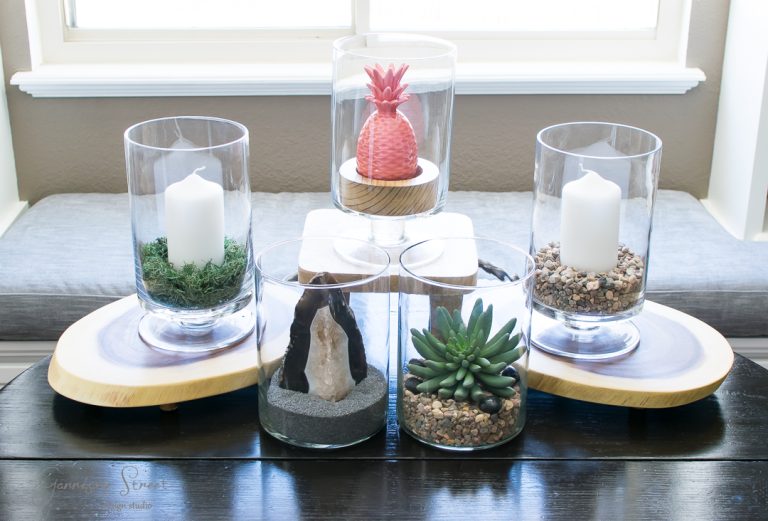 A decorative display with five glass vases arranged on wooden bases. Two vases contain white candles on pebbles, one has a pink pineapple ornament, another displays a green succulent, and the last contains a crystal with sand and stones.