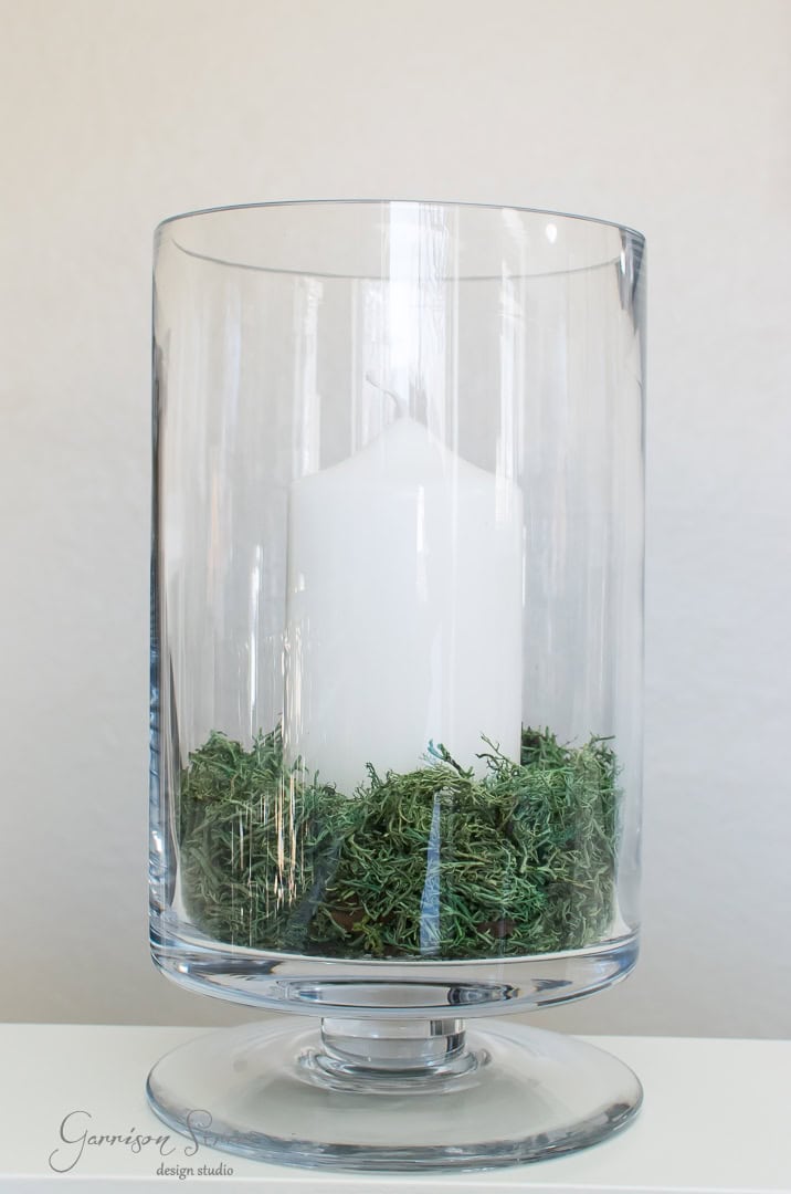 A large white candle sits inside a clear glass hurricane vase, surrounded by green moss. The vase is set against a plain background, creating a minimalist and elegant display.