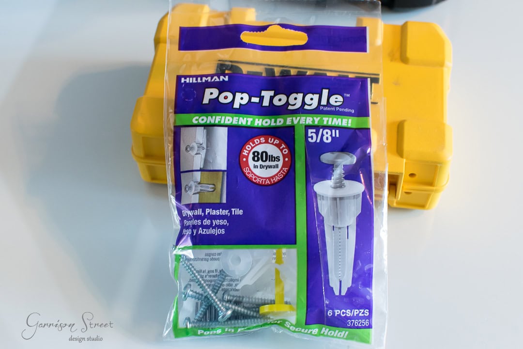 A package of Hillman Pop-Toggle wall anchors, perfect for securing a barn door, is on display. Highlighting a holding strength of up to 80 lbs in drywall, the package includes six pieces with screws. A yellow toolbox sits in the background.