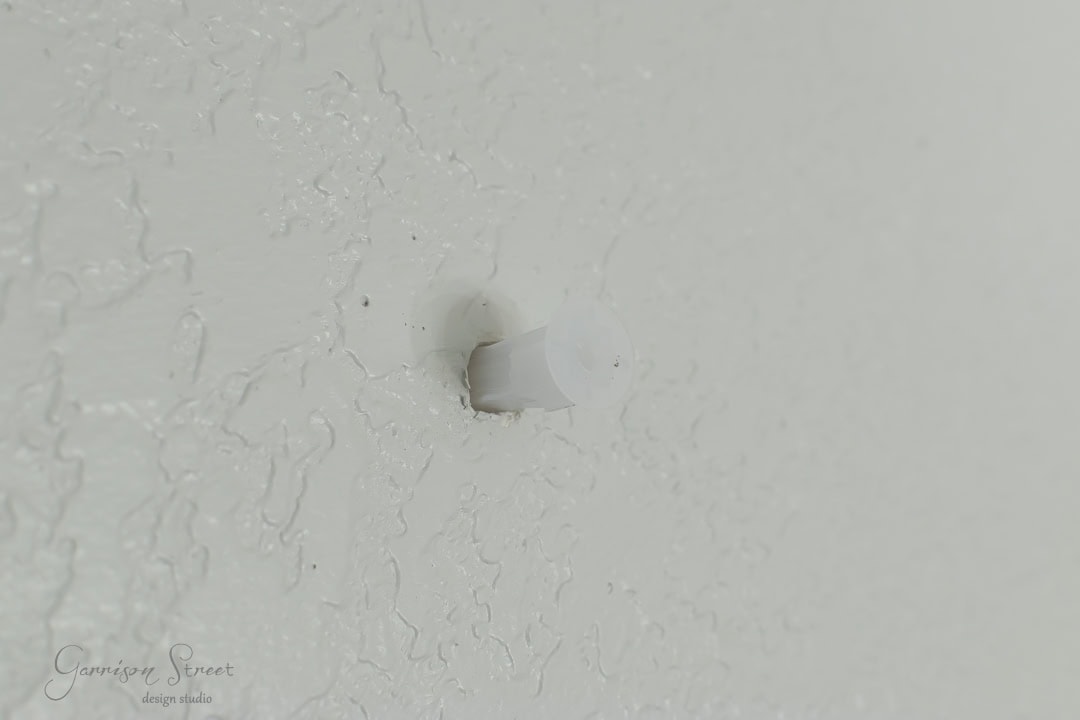 A white plastic anchor is inserted into a textured white wall, designed to hold screws securely. The wall's subtle variations in texture suggest a painted finish, complementing the rustic charm of a nearby barn door.