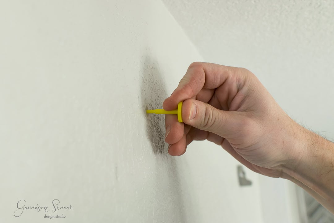 A hand holding a yellow screwdriver is scraping a textured white wall, like the rustic charm of a barn door being brought to life in this DIY project.