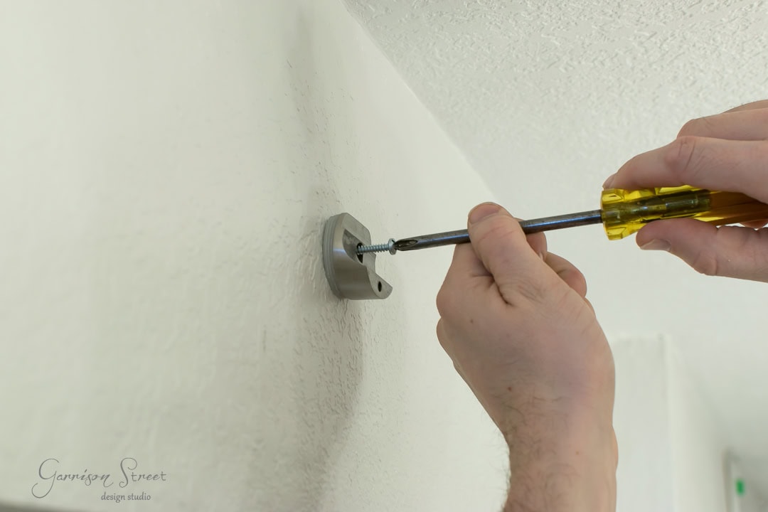 Close-up of a person using a screwdriver to fasten a metal bracket onto a textured white wall, preparing to install a rustic barn door.