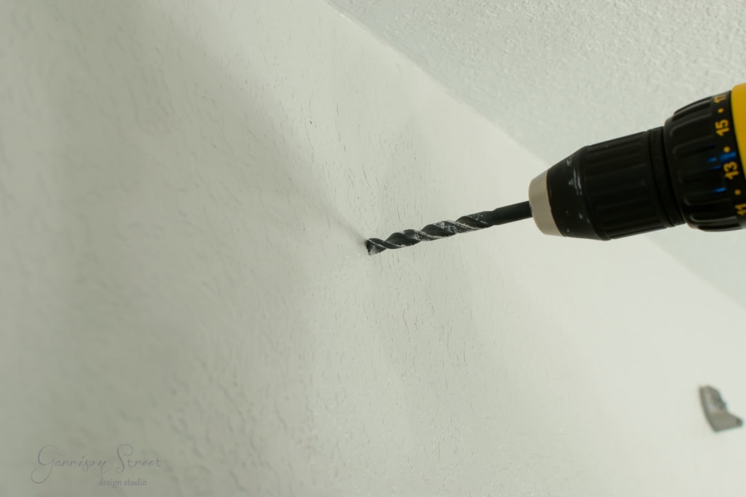 A power drill is being used to drill a hole in a textured white wall, reminiscent of the rustic charm found in barn doors. The drill bit is partially inserted into the wall, and the tool is angled slightly upward.