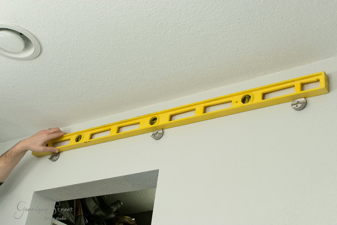 A person measuring a white wall with a yellow level tool, ensuring it's straight. The level is placed above a small opening in the wall, resembling where barn door brackets might fit, and two metallic brackets are visible on the wall.