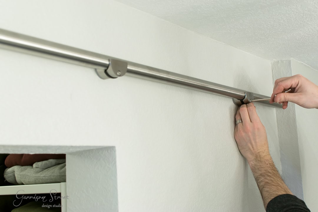 A person adjusts a silver curtain rod mounted on a white wall, reminiscent of the rustic charm of a barn door. Their hands are visible, working near a corner of the room. Below the rod, a small closet with folded clothes is partially visible.